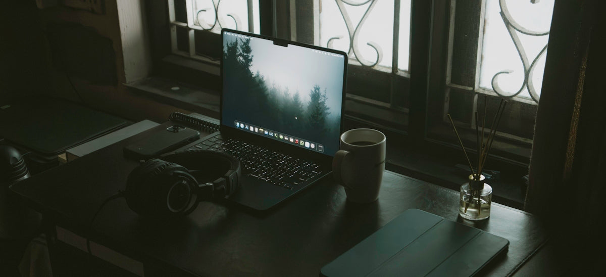 Desk with laptop, headphones, and coffee cup near window.